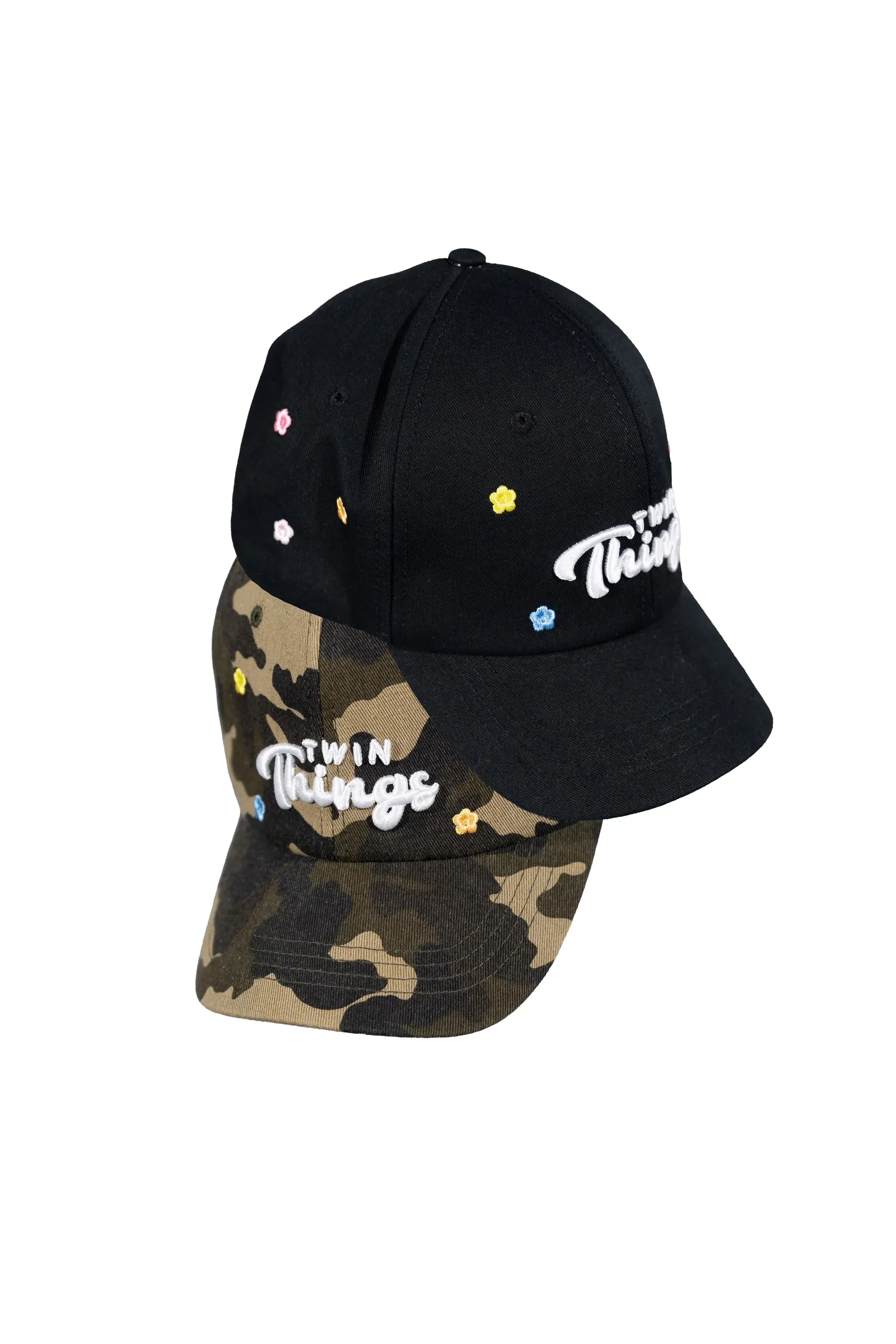Coppia di cappellini baseball Twin Things, uno nero e uno camo, con logo script Twin Things ricamato e microfiori colorati sul fronte
